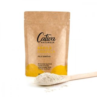 Fundo branco com embalagem de papel ao centro da imagem descrito Cativa Natureza argila amarela, pele sensível, rica em silício fonte minerais selecionadas 100% naturais do Brasil, 100g. Ao lado da embalagem uma colher de madeira cheia com argila amarela.